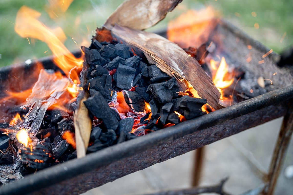 A worn, rusted steel fire box with charcoal and wood burning inside it